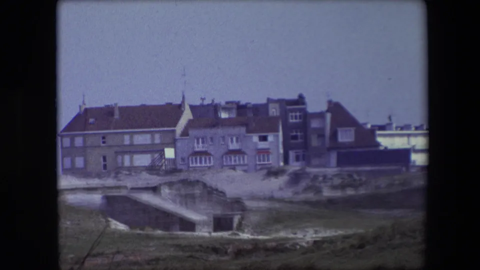 1967 Calais frame with the building shifted to the right of view, showing red roofs, multiple windows, sandy ground, grass, and a low concrete form.