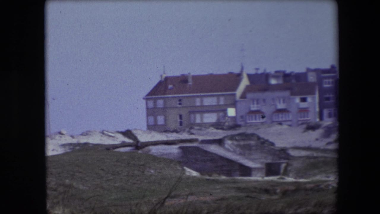 1967 Calais home-movie frame showing grass, pale ground, a concrete drainage structure, and a row of residential buildings in the distance.