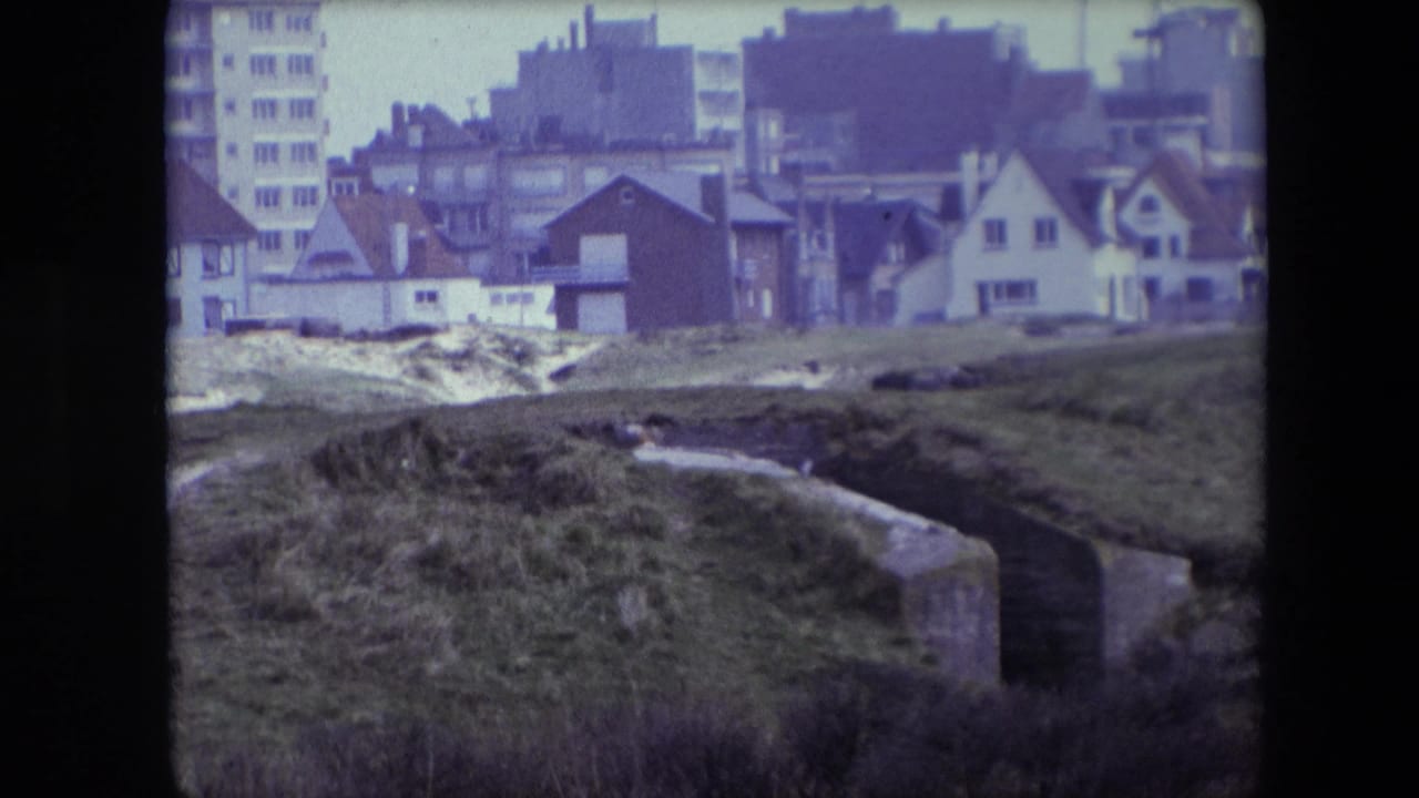 Concrete channel cuts through rough grass with houses and apartment blocks behind.