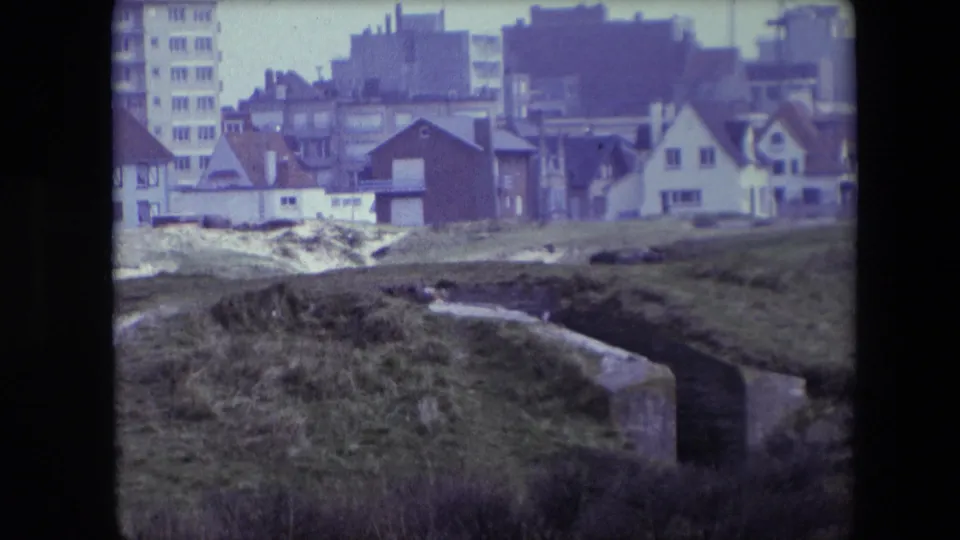 1967 Calais frame looking across rough grass and a concrete drainage channel toward small houses and taller apartment buildings.
