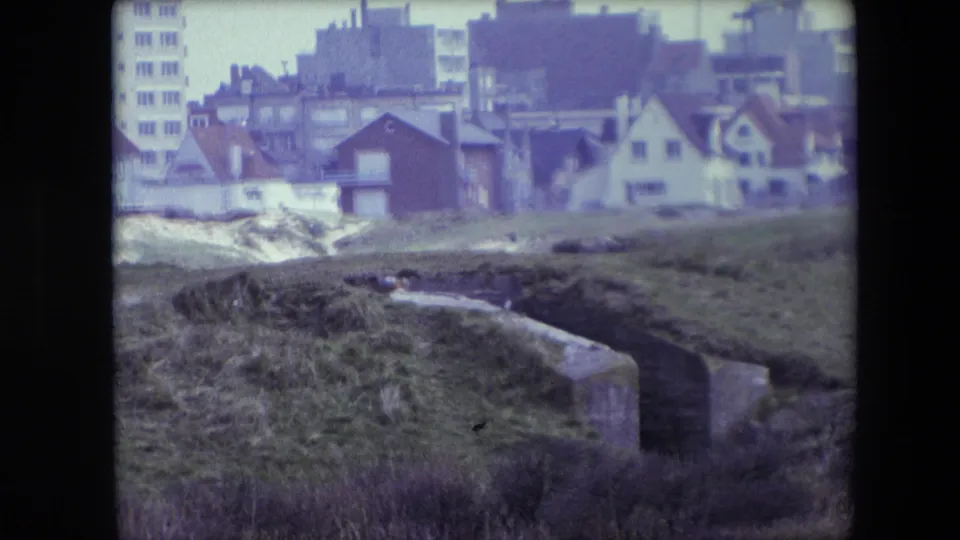 1967 Calais home-movie view of uneven grass, a concrete drainage wall, and clustered houses backed by taller city buildings.