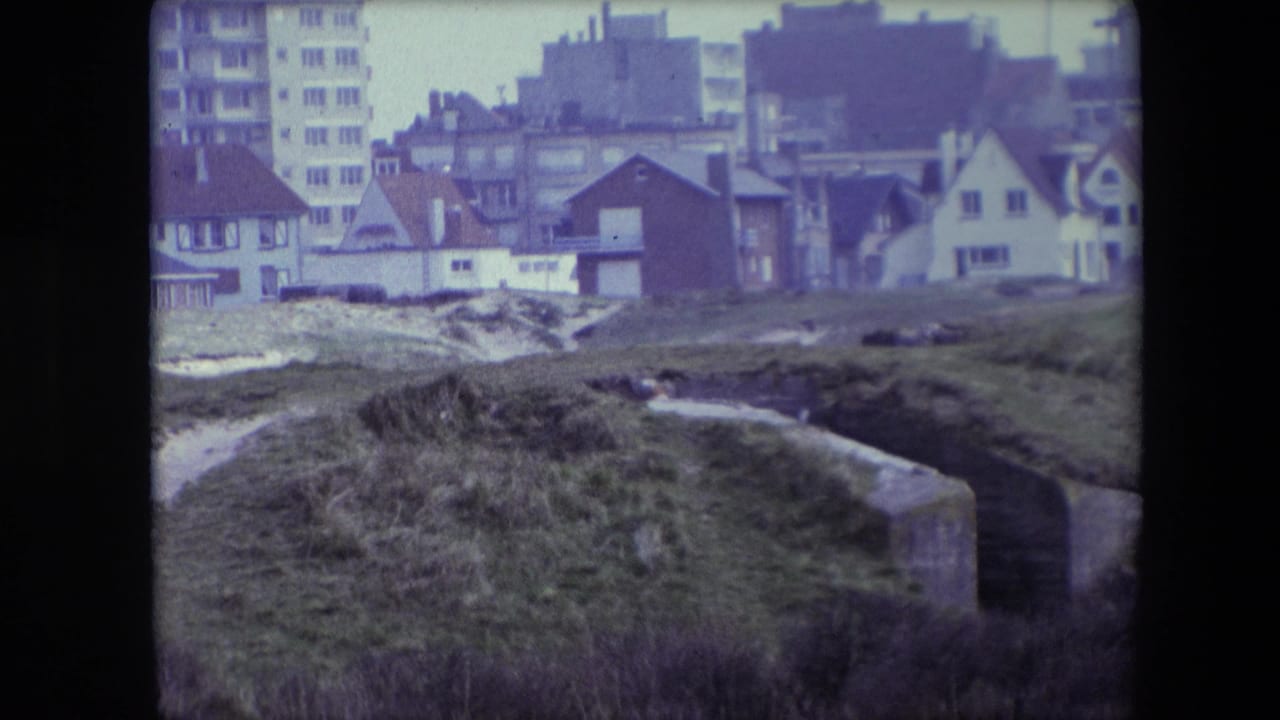 1967 Calais 8mm frame showing grassy dunes and weathered concrete bunker remains with residential houses and a taller apartment block behind them.