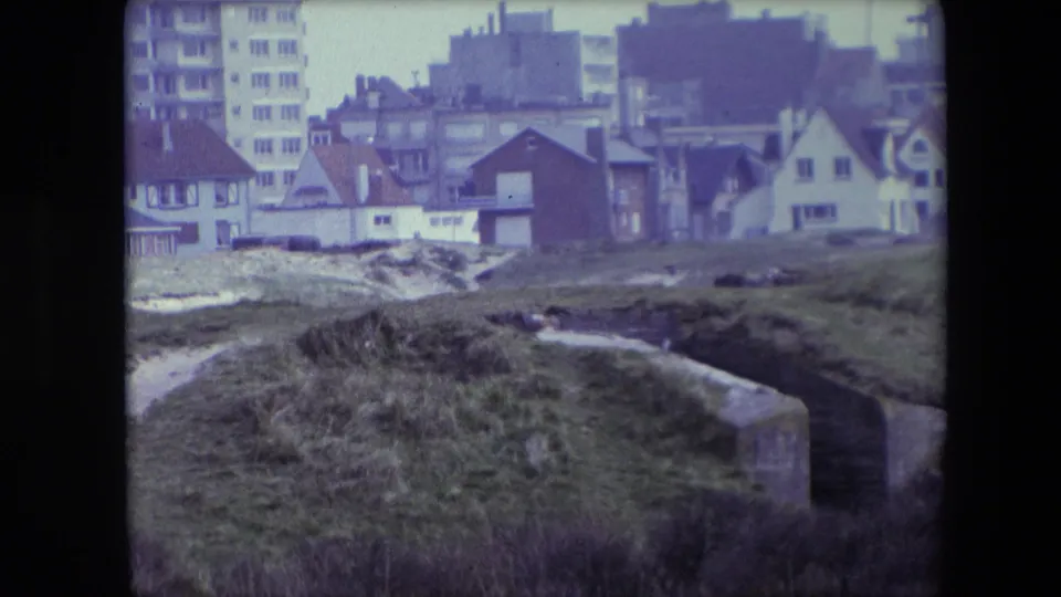 1967 Calais 8mm frame showing grassy dunes and weathered concrete bunker remains with residential houses and a taller apartment block behind them.
