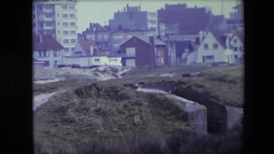 1967 Calais frame with a low concrete bunker or fortification edge in grass, backed by houses and a tall apartment building.