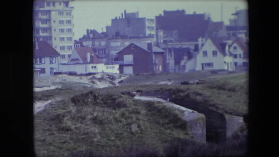 1967 Calais home-movie frame showing grass, pale ground, a concrete bunker edge, houses, and a taller apartment block in the background.