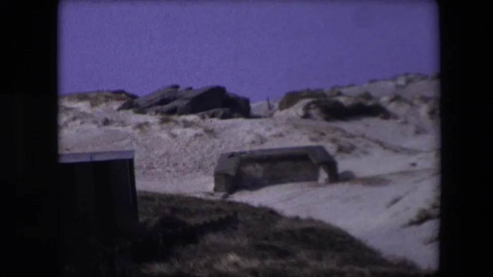 1967 Calais frame showing sandy ground, low grassy mounds, and weathered concrete bunker openings under a purple-tinted sky.