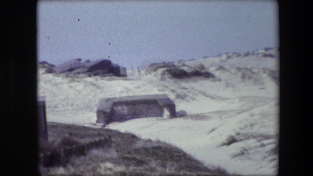 1967 Calais home-movie frame showing pale dunes, dark rocks, scrubby grass, and a low weathered concrete structure partly set into the sand.