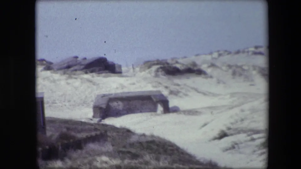 1967 Calais home-movie frame showing pale dunes, dark rocks, scrubby grass, and a low weathered concrete structure partly set into the sand.