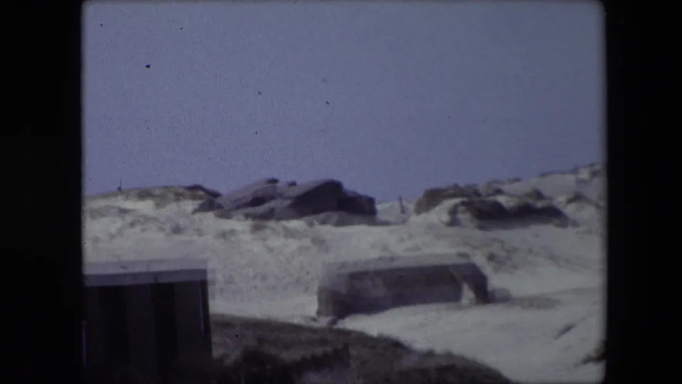 1967 Calais home-movie view of a dune slope, scattered rocks, a partially visible dark building edge, and a low concrete structure in the sand.