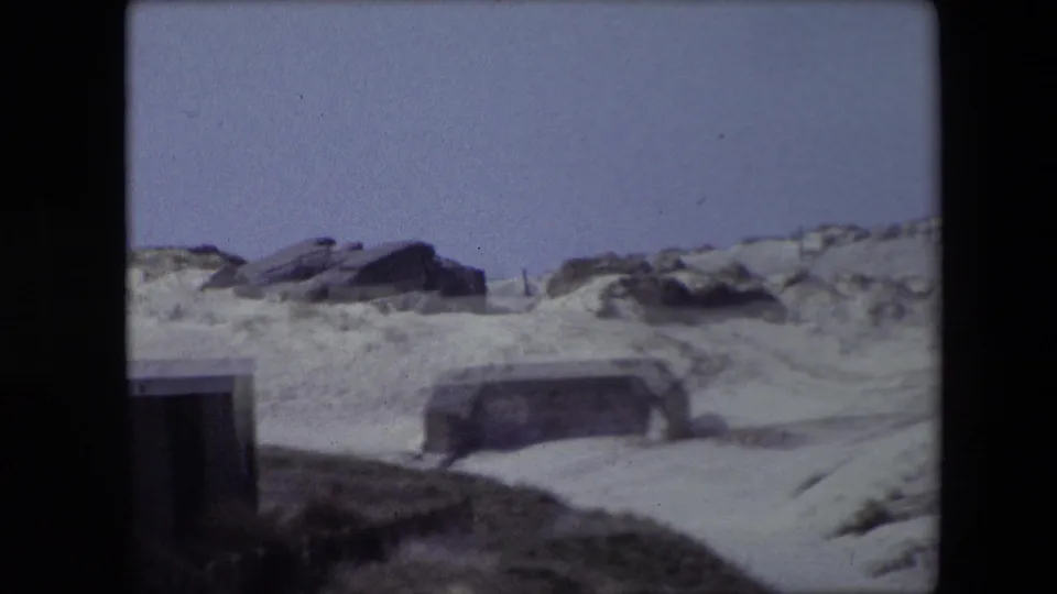 1967 Calais frame showing a quiet sandy coastal scene with a weathered concrete structure below dune ridges, rocks, sparse grass, and blue sky.