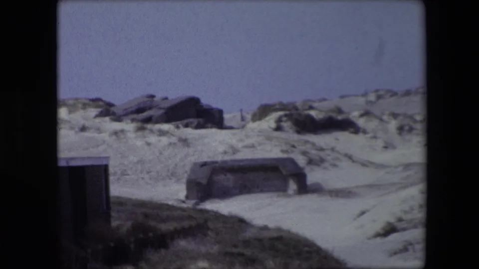 1967 Calais-area 8mm frame showing weathered concrete bunker forms set into sandy coastal dunes beneath a washed-out sky.