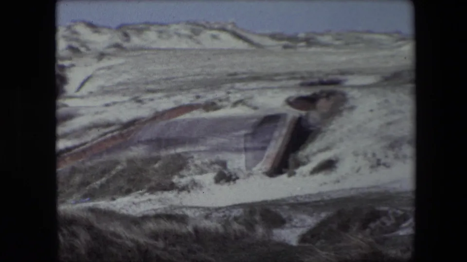 A handheld 8mm view of a concrete bunker partly swallowed by Calais dunes, with grass and pale sand surrounding the exposed structure.