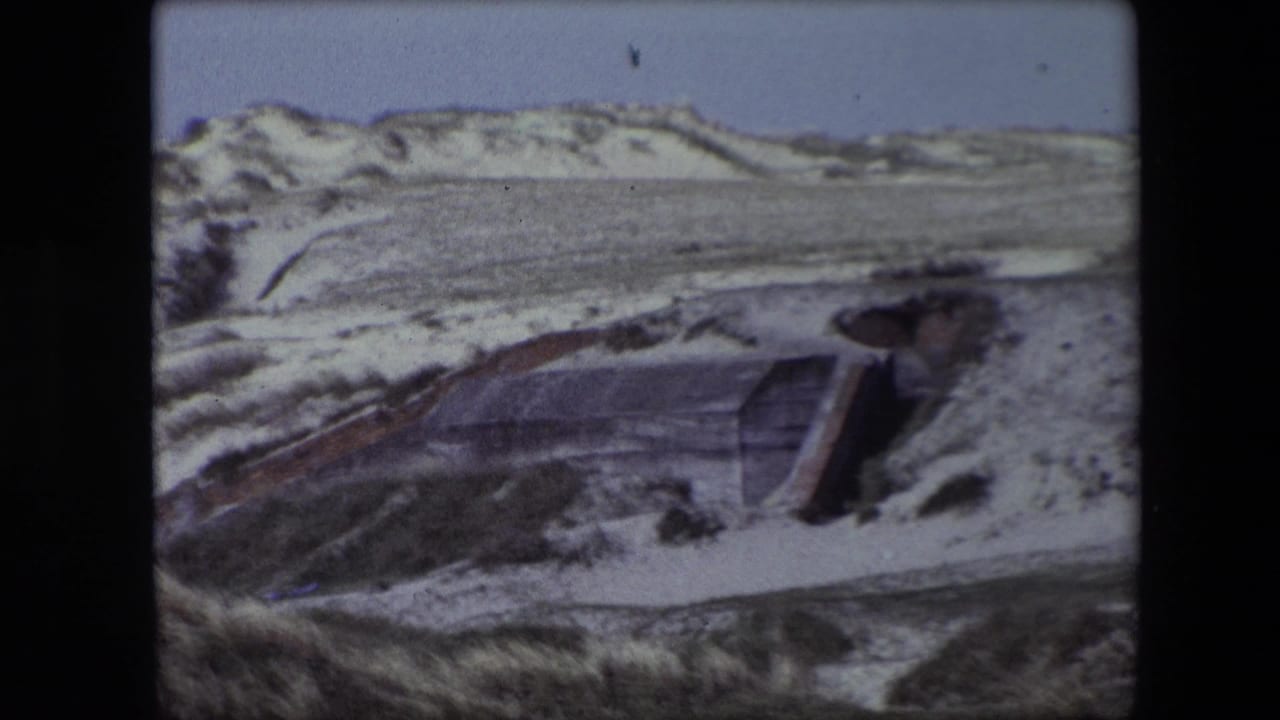 1967 Calais home-movie frame showing pale winter dunes, sparse grasses, and a low weathered concrete structure beneath a clear blue sky.