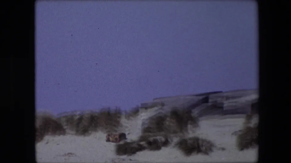 1967 Calais home-movie frame looking across pale dunes and grass clumps toward a low structure under an open blue sky.
