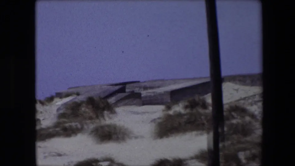1967 Calais frame showing pale dunes, scattered grasses, and a low concrete structure, with a dark vertical edge mark from the film scan.