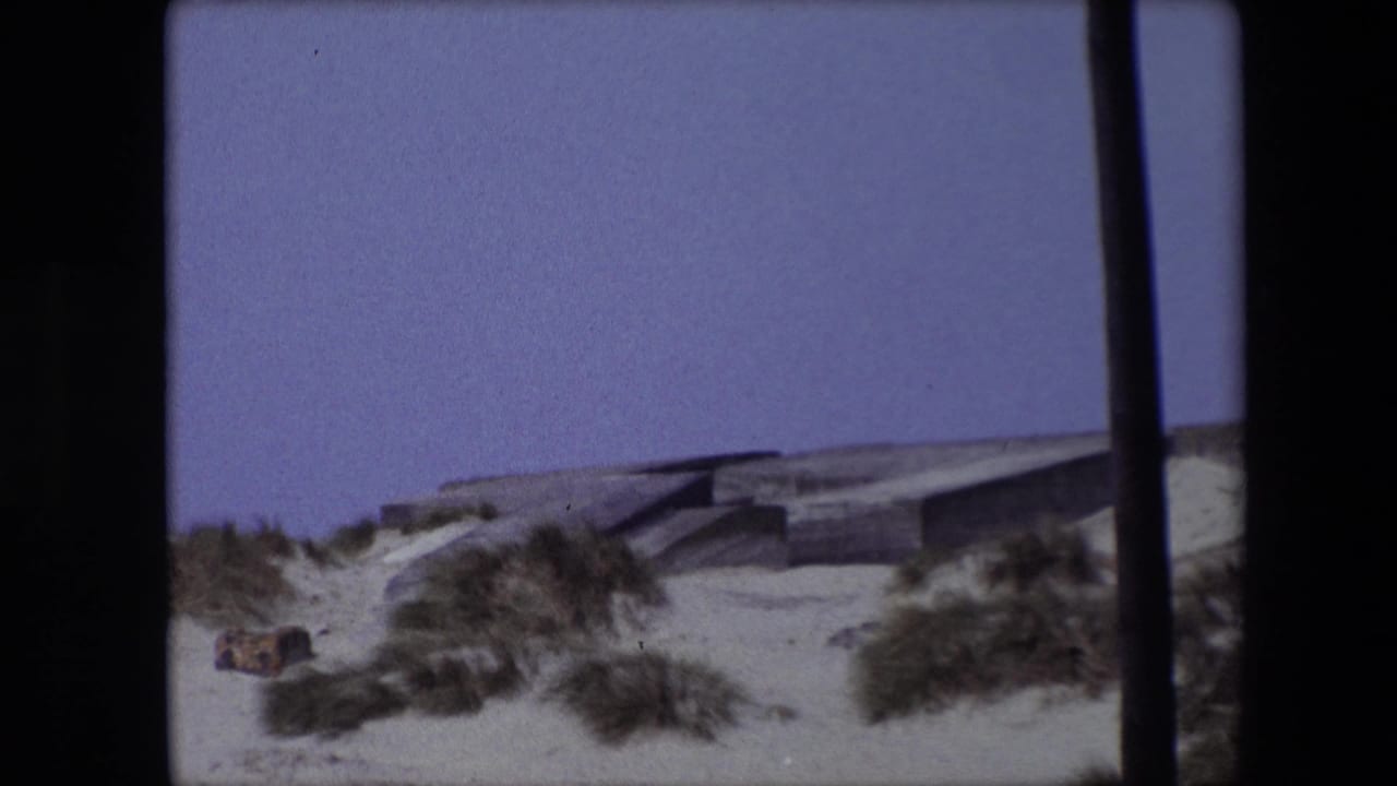 Concrete bunker-like structure partly visible beyond sand dunes and scrub grass under a pale sky.