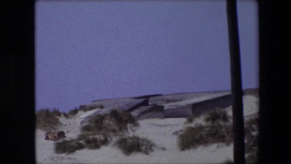 1967 Calais 8mm frame showing a low concrete coastal bunker-like structure behind sandy dunes, with scrub grass and a dark vertical obstruction at the right edge.