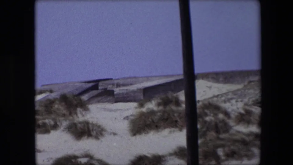 1967 Calais 8mm frame with a concrete bunker-like coastal form set behind pale dunes and dune grass, bordered by dark vertical film or foreground obstructions.