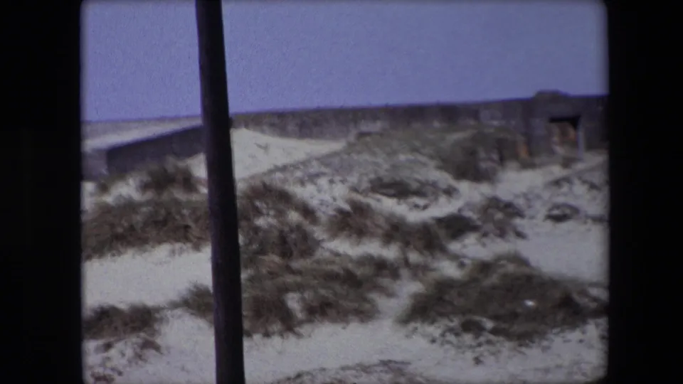 1967 Calais 8mm frame looking across sandy coastal dunes toward a bunker-like concrete structure with a dark opening, partially obscured by a vertical foreground bar.