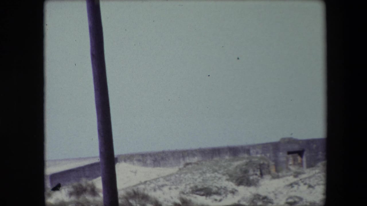 1967 Calais home-movie frame showing pale dunes, scrub vegetation, and a low weathered stone or concrete structure under a washed-out sky, with dark vertical poles in the foreground.