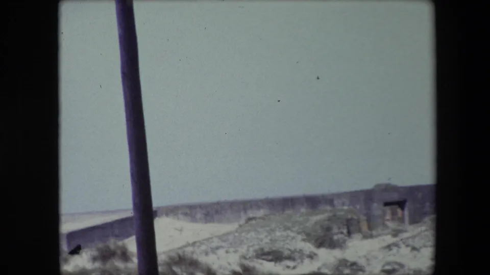 1967 Calais home-movie frame showing pale dunes, scrub vegetation, and a low weathered stone or concrete structure under a washed-out sky, with dark vertical poles in the foreground.