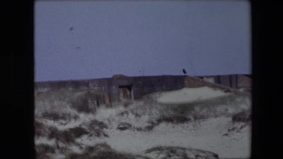 1967 Calais home-movie frame showing a low weathered ruin with a dark opening, pale sand, rough dune vegetation, and a broad muted sky.