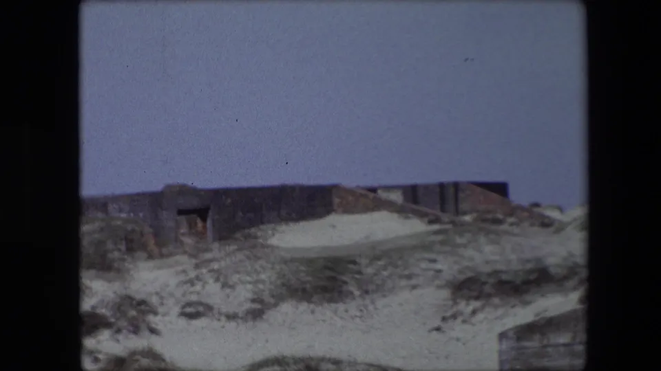 1967 Calais frame showing the low weathered structure continuing across the dunes, with pale sand, sparse grasses, and a small edge of another built element at the lower right.