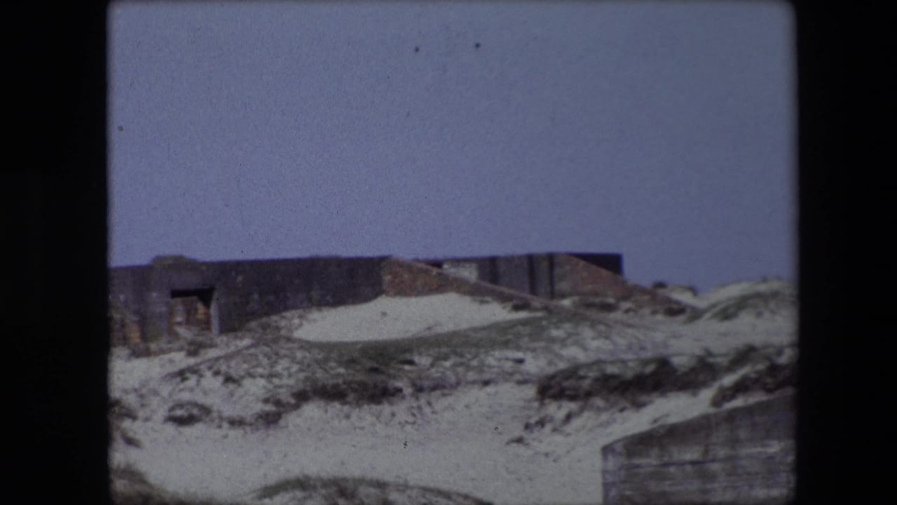 1967 Calais home-movie frame showing sand, scrub grass, and a low weathered structure set into a rockface under a pale blue sky.