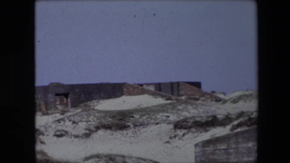 1967 Calais home-movie frame showing sand, scrub grass, and a low weathered structure set into a rockface under a pale blue sky.