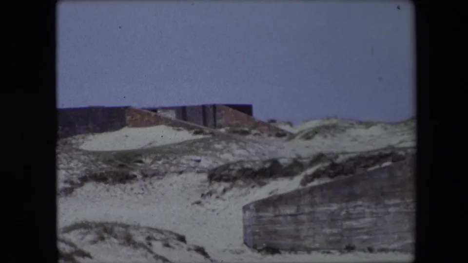 1967 Calais frame with a concrete or stone wall crossing the lower right and sandy dune-like ground rising toward a partial structure above.