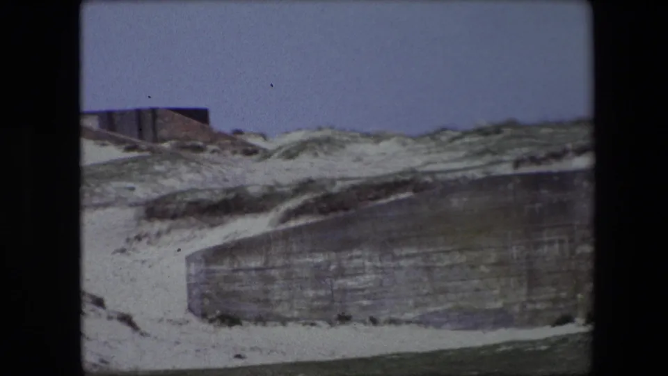 Calais 1967 8mm frame showing a long weathered wall or bunker face below a sandy, grass-covered rockface during a handheld pan.
