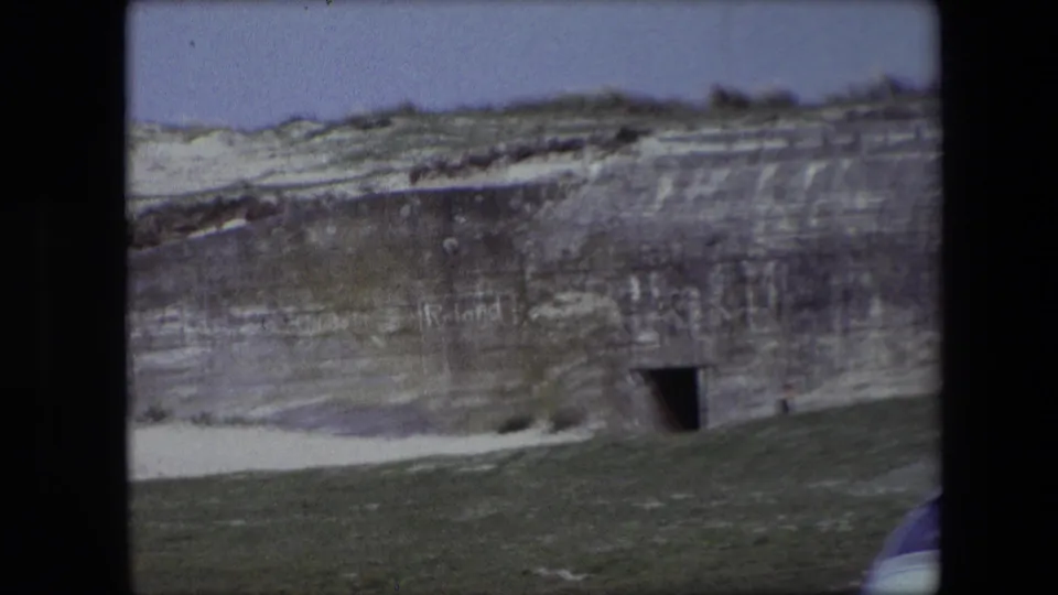 1967 Calais frame showing a weathered wall embedded in a sandy slope, with a dark rectangular opening and faint markings on the concrete surface.