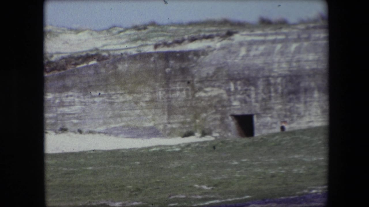 1967 Calais home-movie frame showing a large weathered concrete bunker structure in a grassy slope, with a dark rectangular entrance, pale snow patches, and a small figure near the wall.