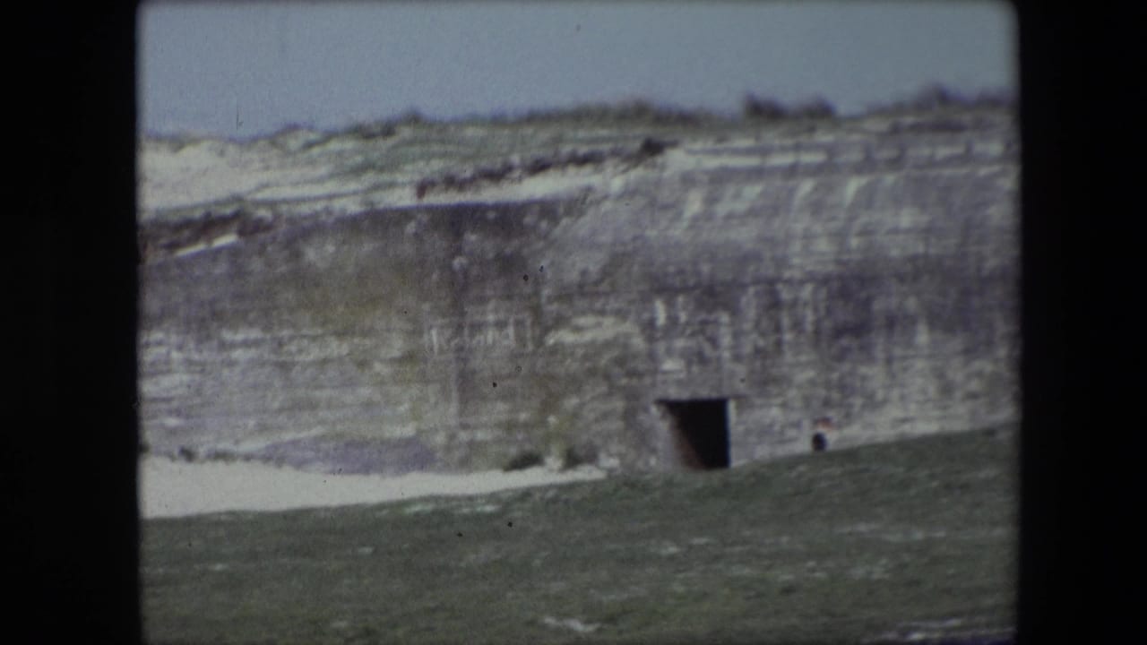 Concrete hillside bunker with a dark opening, sloped grass, pale sky, and a small figure near the wall.