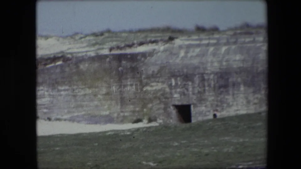 1967 Calais frame of the same weathered concrete bunker set into a grassy hillside, with a dark entrance, snow on the ground, and a tiny person visible near the structure.