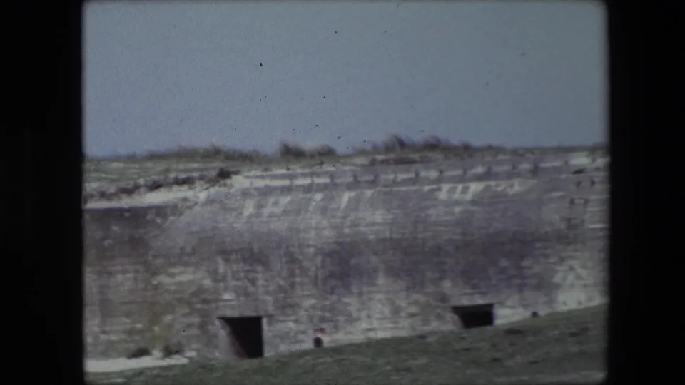 1967 Calais home-movie frame showing a long weathered concrete bunker wall with two dark rectangular openings, a grassy foreground, faint markings, and tiny people near the base.