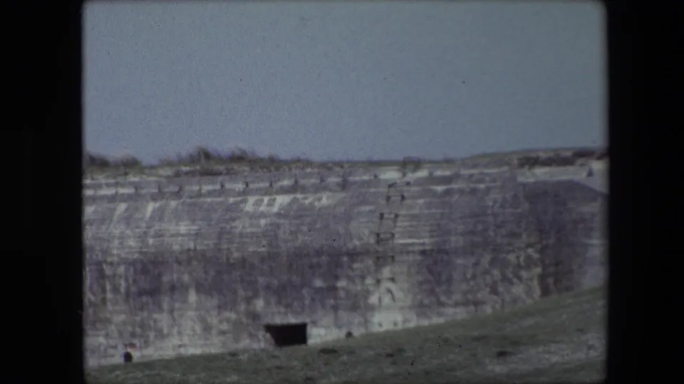Final 1967 Calais frame showing the concrete bunker wall from a slightly shifted handheld angle, with the grassy slope rising across the foreground and a dark opening near the lower wall.