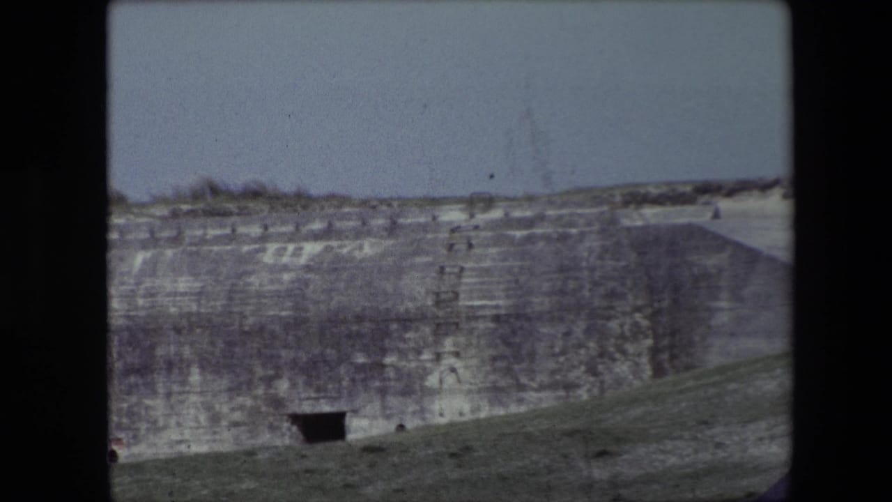 1967 Calais, France 8mm frame showing a pale sky above a broad carved rock face with dark cave-like entrances and a grassy foreground slope.
