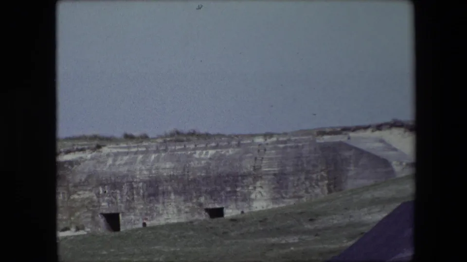 1967 Calais, France frame with a wider view of the excavated hillside, two dark cave-like openings, sparse vegetation, and a sloping foreground.