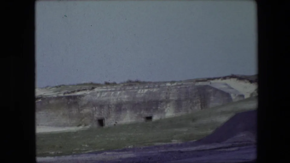1967 Calais, France 8mm frame showing the carved hillside from farther back, with small dark openings, grassy ground, and gravel or dirt in the foreground.
