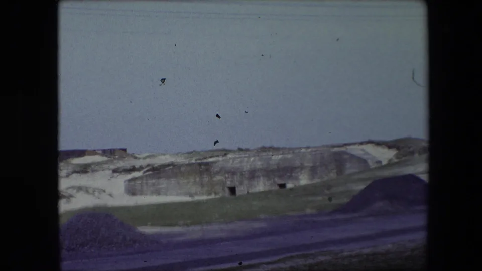 1967 Calais, France frame showing the cave-like hillside at a greater distance, with dark entrances, a pale sky, grassy slope, and a dark mound or foreground form.