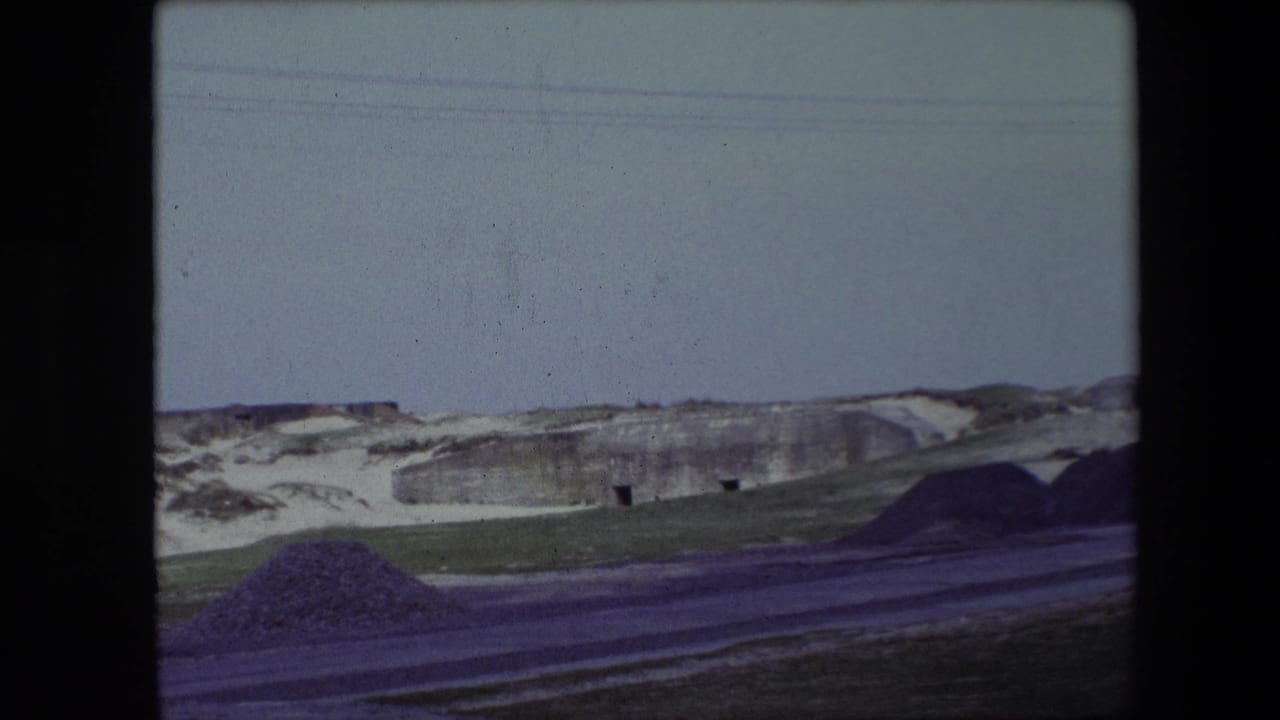1967 Calais home-movie frame showing snow-covered low hills, a roadside, grass, and a weathered concrete structure beneath a pale winter sky.