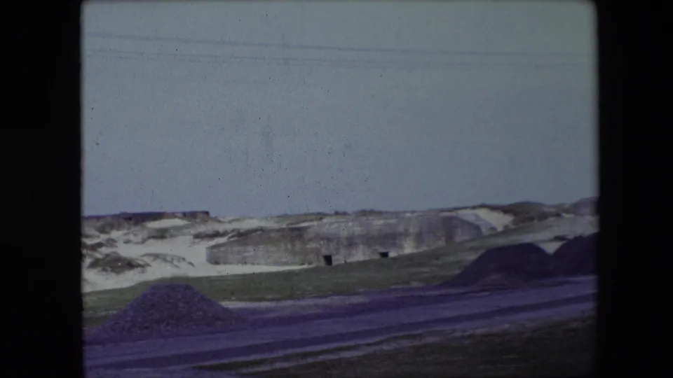 1967 Calais home-movie frame showing snow-covered low hills, a roadside, grass, and a weathered concrete structure beneath a pale winter sky.