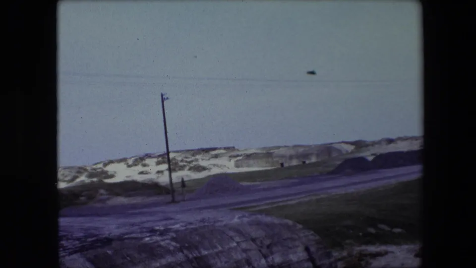 1967 Calais 8mm frame showing a winter roadside scene with utility poles, overhead lines, snow patches, grass, and a low concrete structure.