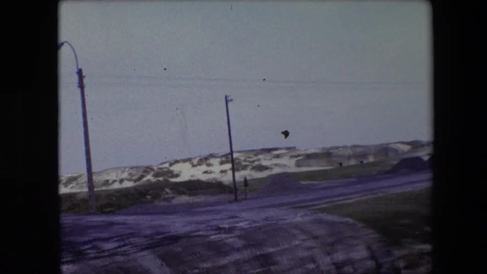1967 Calais frame showing a snowy roadside, utility poles, overhead lines, distant concrete structure, and a small bird crossing the pale sky.