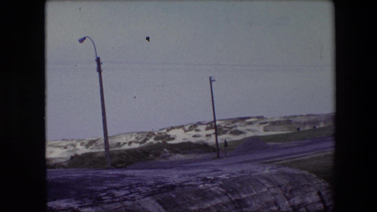 1967 Calais, France home-movie frame showing a quiet snow-dusted road curving past sandy dunes, with a curved streetlight and utility poles against a pale sky.