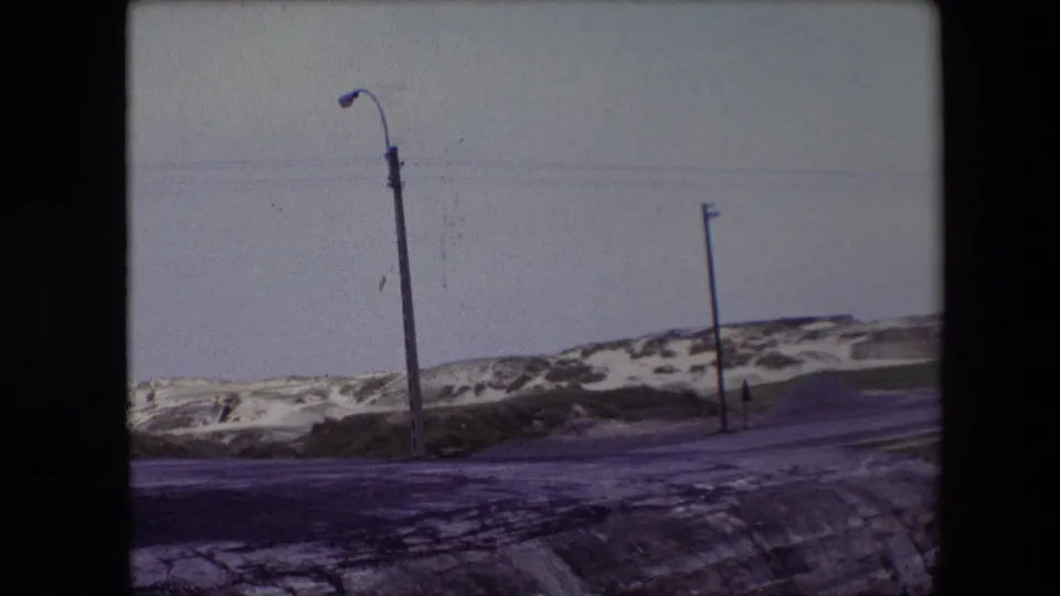 1967 Calais, France frame of a quiet roadside dune area, with snow patches along the asphalt, a curved streetlight, a utility pole, and muted winter light.