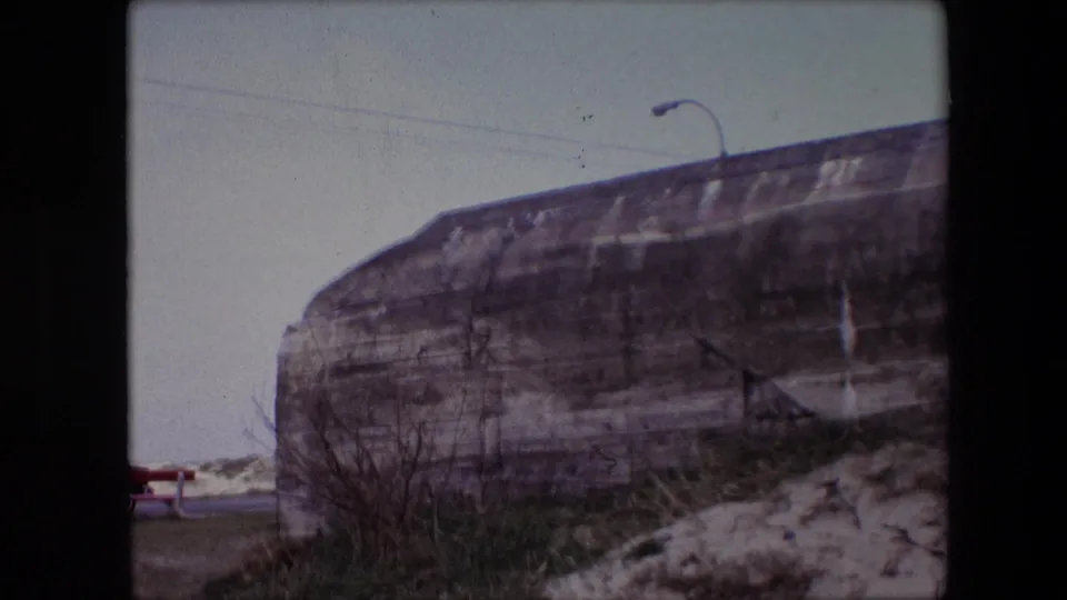 1967 Calais, France home-movie frame showing a weathered concrete roadside structure beside low brush and snow, with a streetlight rising behind it.