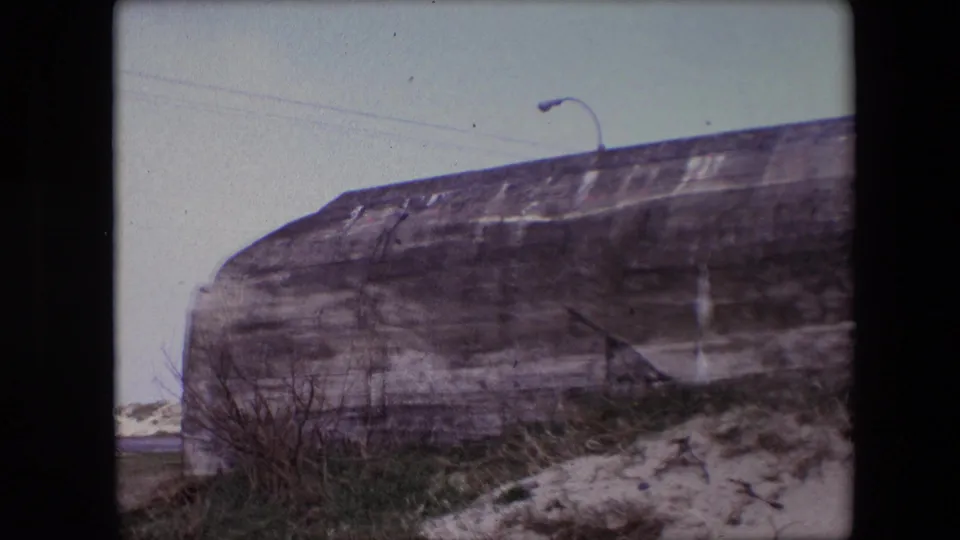 1967 Calais, France frame of a weathered concrete roadside structure filling the foreground, with scrub vegetation, snow patches, and a partially visible streetlight above.