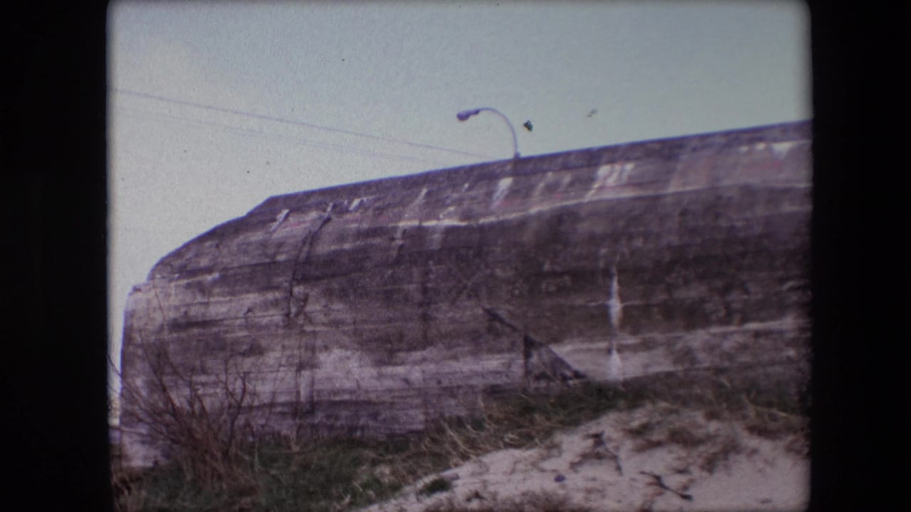 1967 Calais 8mm frame showing a weathered concrete bunker wall, sandy foreground, sparse grass, and a curved streetlamp rising above the top edge.